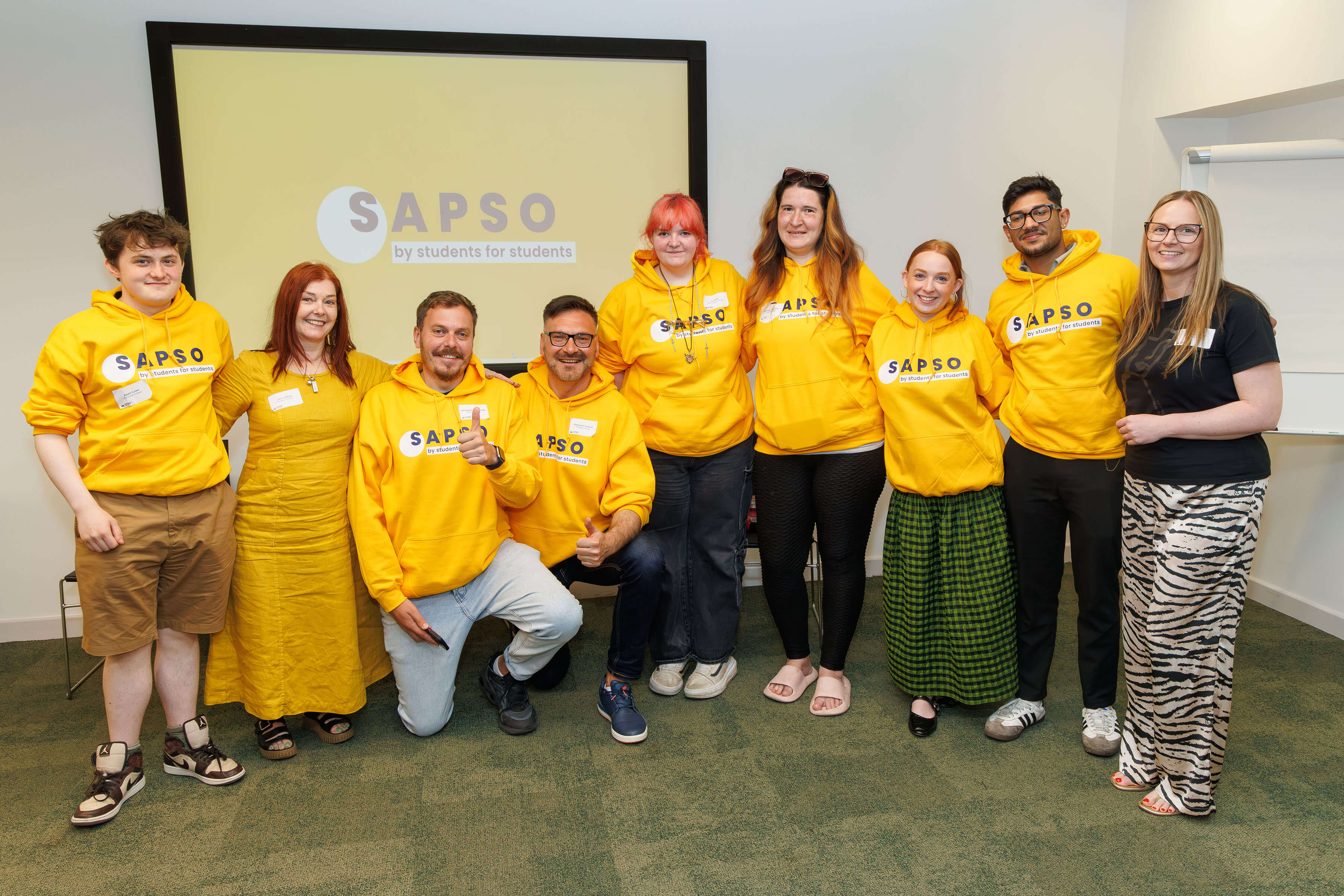 A photo of a project team of nine people standing in front of a projector screen displaying their project logo. Most of them are wearing jumpers with the project logo.