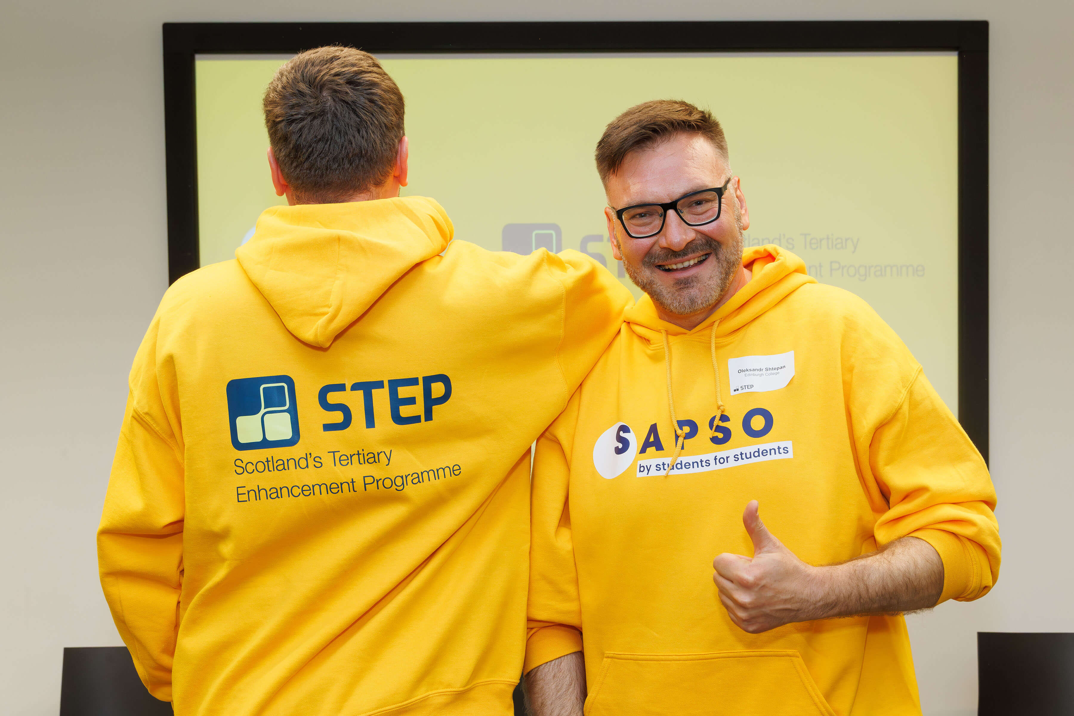 Two students, Volo and Olex, standing in front of a projector screen, modelling their yellow SAPSO hoodies